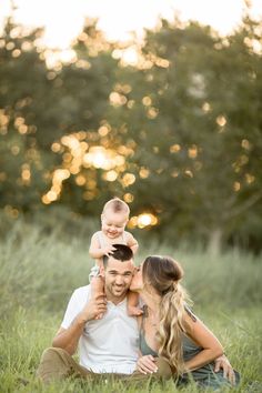 a man and woman are sitting in the grass with their baby on their shoulders while they look at each other