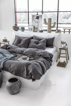 a bed with grey linens and pillows in a white room next to large windows