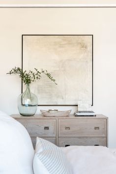 a vase with some flowers sitting on top of a dresser next to a painting and pillows