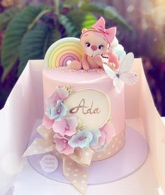 a small pink cake with a teddy bear on top and flowers around the edges, sitting in a box