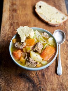 a bowl of soup with meat, carrots and bread on a wooden cutting board