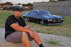 a man sitting on top of a cement slab next to a black sports car in the background