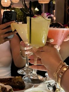 two women holding up drinks at a table with food and drink glasses on it, while another woman takes a photo