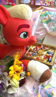 a stuffed animal next to a box of candy and flowers on a bed with sheets