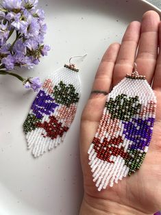 a hand holding a pair of beaded earrings on top of a white plate next to purple flowers