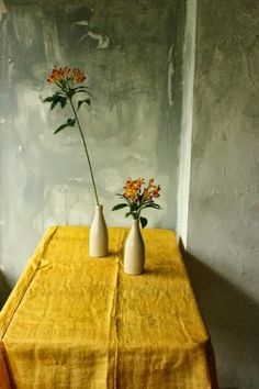 two white vases with flowers in them on a yellow linen tablecloth against a gray wall