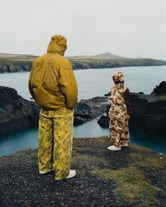 two people standing on the edge of a cliff overlooking water and land, with one person in yellow jacket looking at another