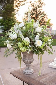 a vase filled with white and green flowers on top of a wooden table next to trees