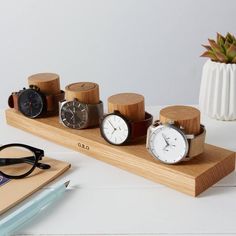 five different watches on a wooden stand next to a pen and eyeglasses, with a plant in the background