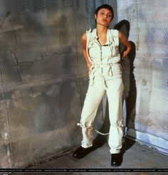 a woman standing in front of a fence with her hands on her hips and wearing white overalls