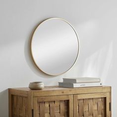 a round mirror on top of a wooden cabinet next to a bowl and bookcase