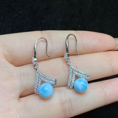 "This pair of 7mm Larimar dangling earrings shows extraordinary charm with its unique flavor and exquisite craftsmanship. They are pristine and bright in silver-colored metal. The focal point of each earring is centered on the carefully selected 7mm Larimar pendant. This Larimar, with its unique texture and color, seems to carry the depth and vastness of the ocean. The blue tone intertwined with white texture, like the delicate traces left by the waves lapping on the beach, and like the morning sun shining on the surface of the sea in the layers of light, both mysterious and fascinating. Surrounding the Larimar is a ring of fine white zircons. These zirconias are small, but they are bright, and they not only add sparkle to the earrings, but also form a harmonious match with the blue color Larimar Dangle Earrings For Gifts, Silver Larimar Dangle Earrings, Elegant Larimar Dangle Earrings, Silver Larimar Dangle Jewelry, Larimar Dangle Earrings As Gift, Jewelry Knowledge, Bridal Earrings Drop, Sun Shining, Larimar Pendant