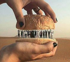 a hand holding a hamburger in the desert with people standing around it on top of it