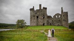 a bride and groom standing in front of an old castle on a grassy hill next to a body of water