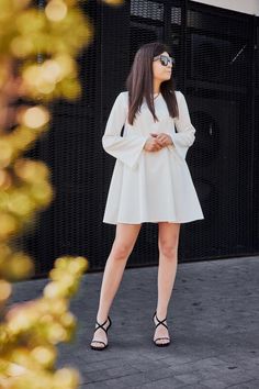 a woman standing in front of a building wearing sunglasses and a white dress with long sleeves