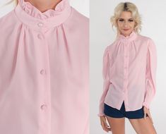 "Vintage 80s blouse in pink with a high neck and ruffled trim. Buttons up the front with long sleeves.  Please see measurements and condition below. Every garment we sell is authentic vintage! You will receive the exact item photographed. Condition: Very good vintage. Best fits women's: Medium Material:  Polyester MEASUREMENTS Taken from seam to seam while the garment is lying flat. Double the armpit, waist, and hips For reference, model is 5'7\" and measures 31-23-35. Length from Top: 24\" Armp Retro Pink Top With Ruffles, Pink Ruffled Collar Blouse For Fall, Retro Pink Ruffled Top, Pink Fitted Top With Ruffled Collar, Pink Collared Top With Ruffles, Fitted Pink Blouse With Ruffled Collar, Pink Button-up Blouse With Ruffles, Pink Ruffled Button-up Blouse, Victorian Blouse