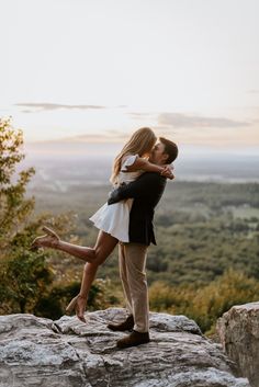 a man and woman are kissing on top of a rock in the mountains at sunset