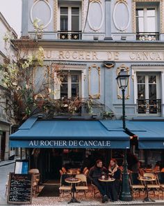 two people sitting at an outdoor cafe with blue awnings in front of the building