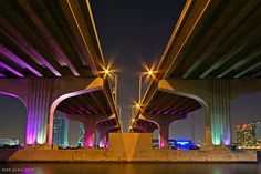 the underside of an overpass at night with lights on and buildings in the background