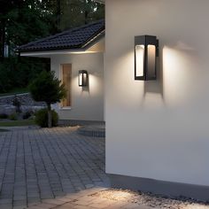 two lights on the side of a white wall next to a brick walkway and trees