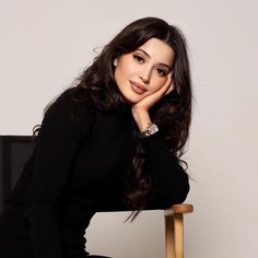 a woman sitting on top of a wooden chair next to a white wall with her hand under her chin