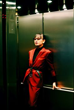 a woman in a red suit leaning against a metal wall with her hand on the door