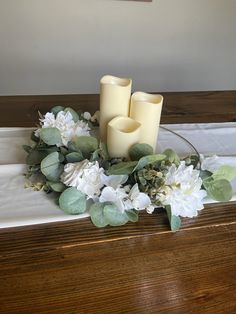 two candles sitting on top of a wooden table next to white flowers and greenery