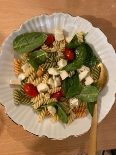 a pasta salad with spinach and tomatoes in a white bowl on a wooden table