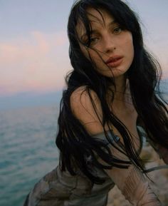 a woman with long black hair standing by the ocean