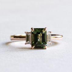 Emerald cut green tourmaline adorned with baguette diamonds Metal: 14K rose gold Stone: Green Tourmaline Stone Size: 8 x6 mm Stone Weight: 1.7 Carat Accent Stone: Baguette Diamonds: Diamond Size: 4.5 x 2 mm (2) Diamond Color and Clarity: GHI, SIDiamond weight: 0.12 carat each Total Diamond Weight: 0.25 carat FREE SHIPP Emerald Gemstone Engagement Ring Gold, Tourmaline And Diamond Ring, Wedding Ring With Emerald Accents, Engagement Rings Emerald Cut Three Stone, Vintage Green Wedding Ring, Green Gem Rings, Green Emerald Engagement Rings, Green Emerald Cut Engagement Ring, Emerald Cut Gemstone Ring