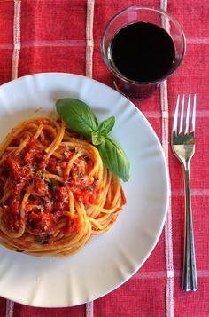 a white plate topped with spaghetti and sauce next to a glass of wine on top of a red table cloth