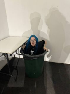 a woman with blue hair sitting in a green trash can next to a white table