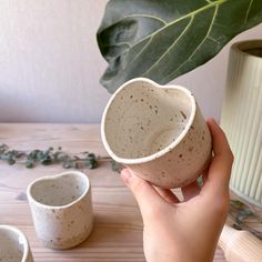 a hand holding a white cup on top of a wooden table next to two cups