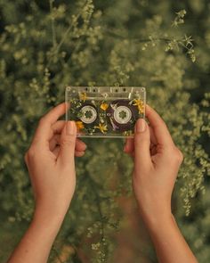 two hands holding up a cassette with eyes and flowers on it in front of trees