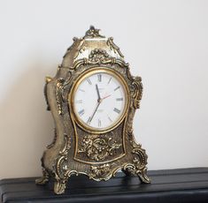 an ornate gold clock sitting on top of a black shelf next to a white wall