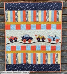 a quilted wall hanging on the side of a wooden fence with cars and trucks