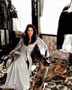 a woman in white dress sitting next to a dog on carpeted floor with clothes