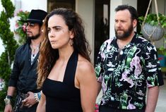 three people standing in front of a house, one woman is wearing a black dress