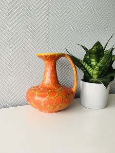 an orange vase sitting next to a potted plant on a white counter top in front of a textured wall