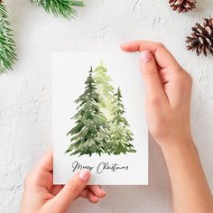 someone holding up a card with a watercolor christmas tree on it and pine cones in the background