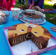 a birthday cake with a lion face cut in half on a blue tablecloth and people sitting around it