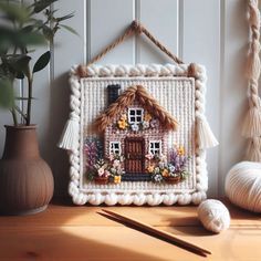 a white frame with a house on it next to some yarn and a potted plant