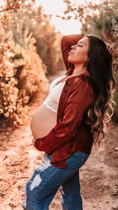 a pregnant woman in jeans and a red hoodie poses for the camera while standing on a dirt path