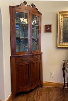 a wooden china cabinet sitting in the corner of a room next to a painting on the wall