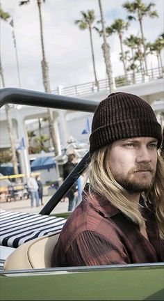 a man with long hair and a beanie sitting in a green convertible car near palm trees