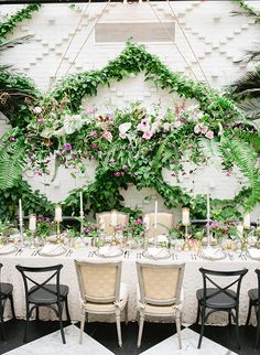 a table set up with white linens and greenery on the wall behind it