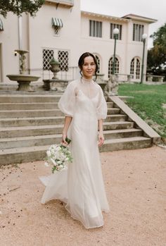 a woman standing in front of a building holding a bouquet and wearing a wedding dress