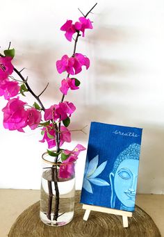 a vase filled with purple flowers next to a small painting on a wooden table top