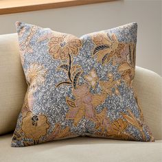 a blue and gold floral pillow sitting on top of a beige couch next to a window