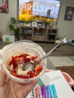 a person holding a plastic cup with strawberries and ice cream in it on top of a table